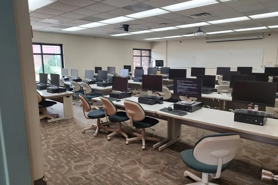 An empty university computer lab with rows of desks, black monitors and office chairs under fluorescent ceiling lights