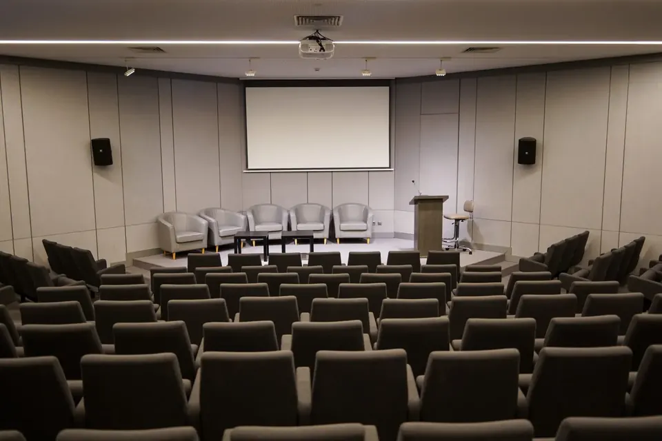 An empty conference room with rows of grey seats facing a low stage with armchairs, a lectern and a blank projection screen