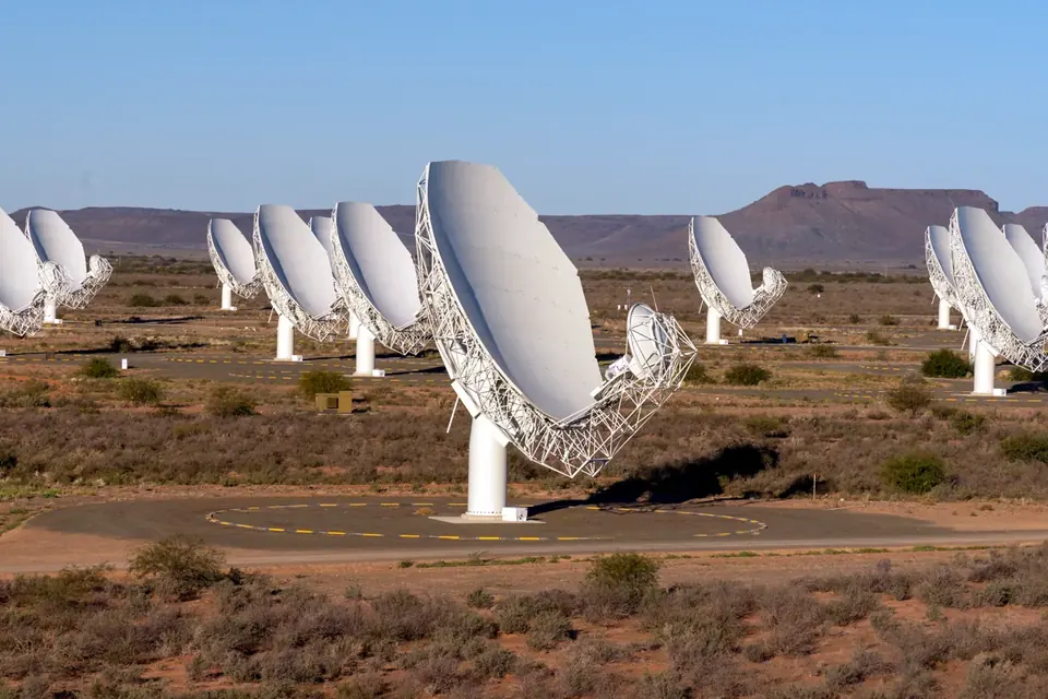 Rows of white radio telescope dishes standing in semi-arid scrubland under a clear sky, with flat-topped hills behind