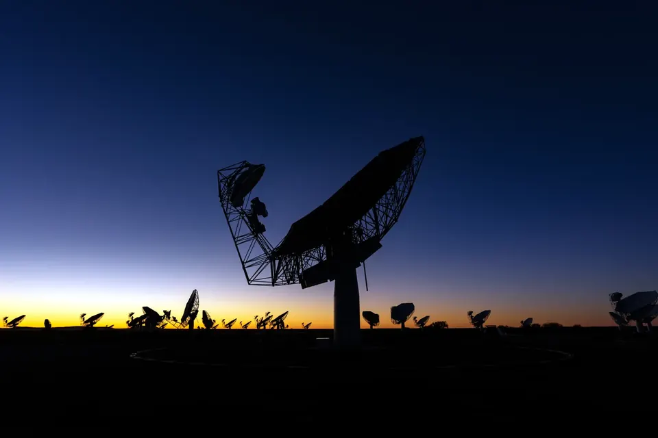 Silhouette of a radio telescope dish against a dark blue evening sky, with dozens of other dishes on the horizon in the Karoo, South Africa