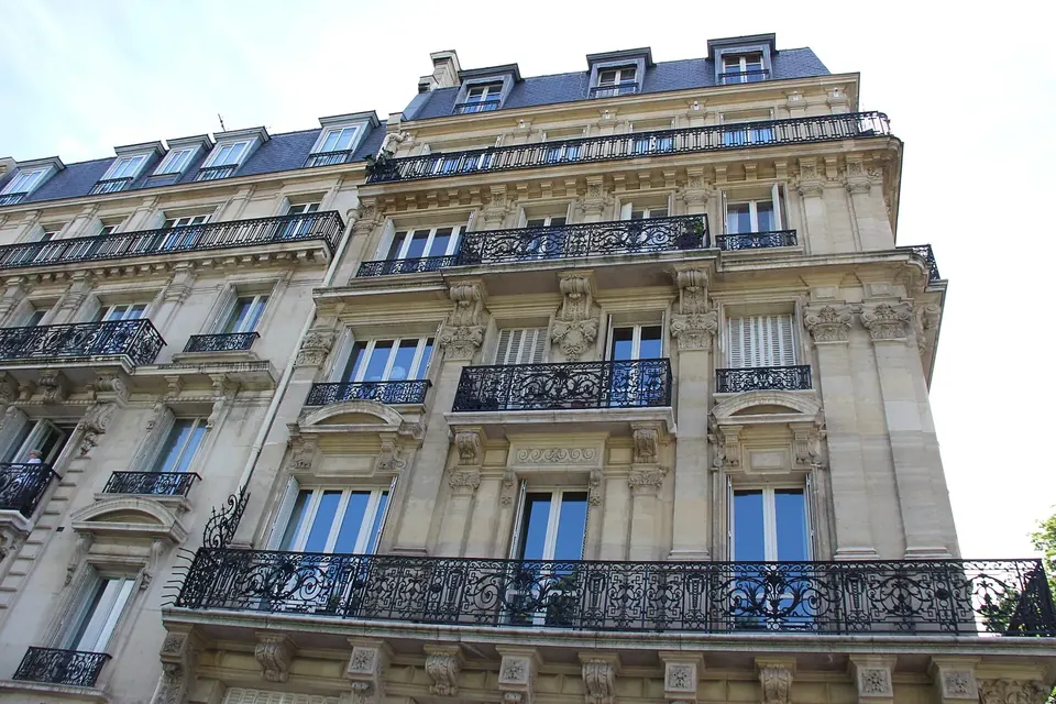 Looking up at the stone facades of two Parisian apartment buildings with wrought-iron balconies, tall windows and grey slate mansard roofs