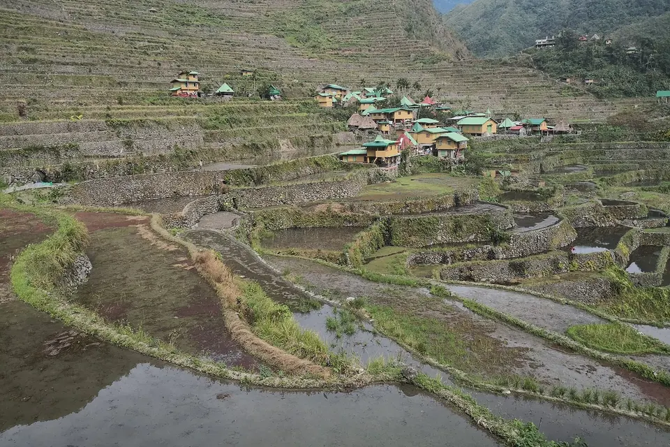 Terraced rice paddies, some flooded, climbing a steep green valley towards a village of small houses under a cloudy sky