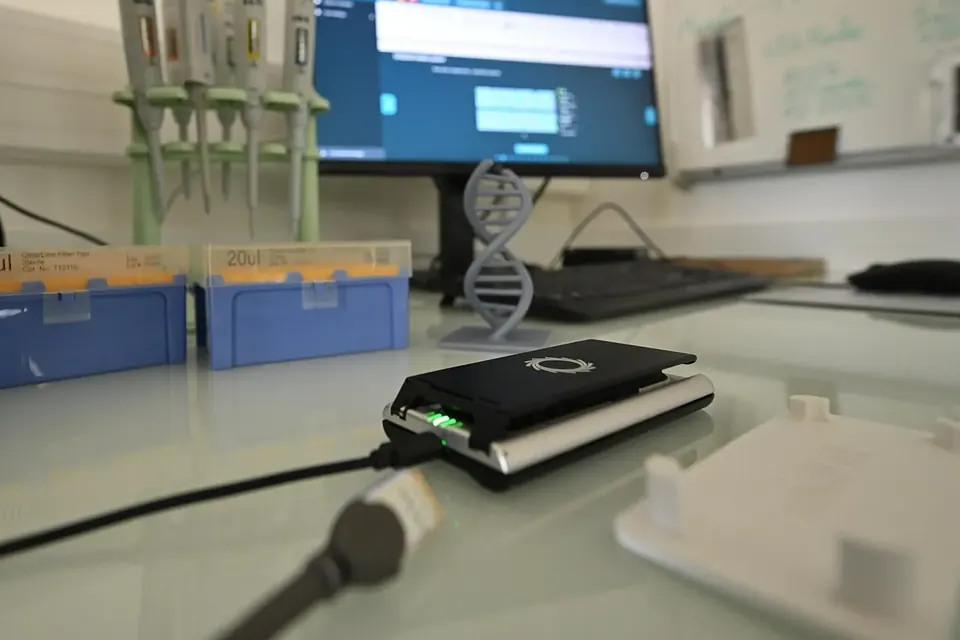 A small portable DNA sequencer connected by cable on a laboratory bench, with pipettes, tip boxes, a model DNA helix and a monitor